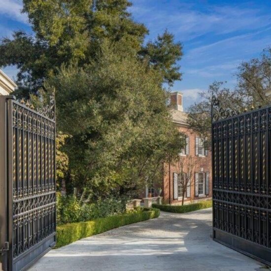 Open black iron gates leading to a tree-lined driveway and a large brick house.